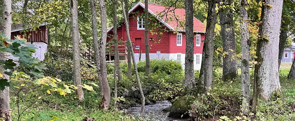 En röd kvarnbyggnad och en bäck. Runt om finns träd och buskar.